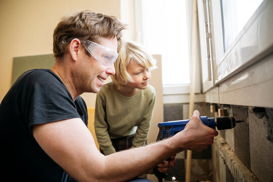 Smiling man drilling wall near son at new home