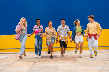 Multi-ethnic group of young friend running in front of yellow wall holding hands