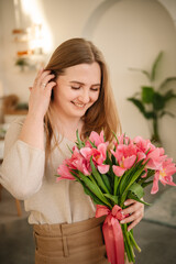 International Women's Day. Beautiful woman is smelling a bunch of spring flowers and standing in living room at home. Portrait young girl in dress holding in hands a big bouquet of pink tulips.