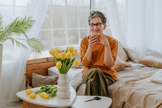 Smiling woman drinking tea near flowers in bedroom at home