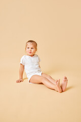 Joyful little baby, pretty girl in white romper sitting on floor with cute cheerful smile against beige studio background. Concept of childhood, motherhood, life, birth. Copy space for ad