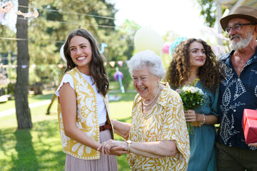 Beautiful senior birthday woman receiving gift from granddaughter. Two mature sisters with grandparents at party. Multigenerational family.