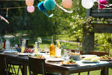 Close up shot of set table at summer garden party, grilled food. Table setting with glasses, lemonade, delicate floral and paper decoration, and bottles of summer wine.