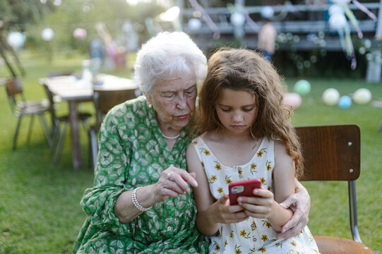 Young girl showing something on smartphone to elderly grandmother at garden party. Love and closeness between grandparent and grandchild.