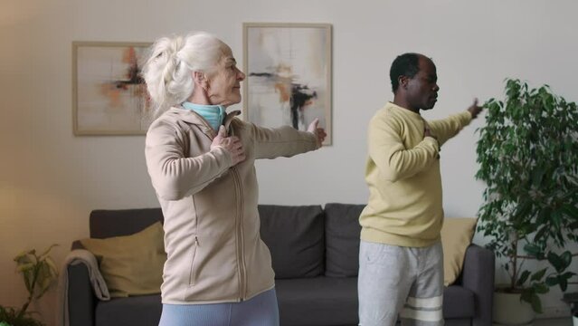 Medium shot of active ethnically diverse couple of seniors doing physical exercises during home workout at daytime