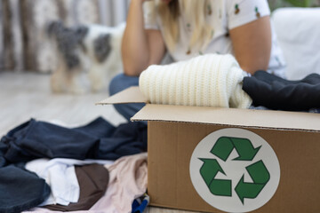 A box with a recycling sign with folded clothes stands on the floor. A woman sits in the background...