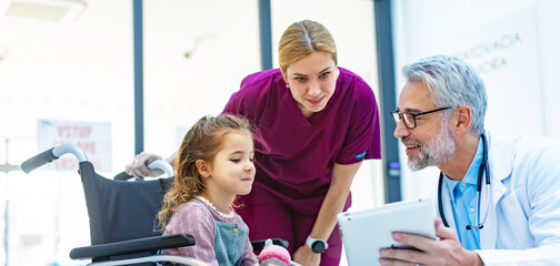 Obraz premium Friendly pediatrician showing someting on tablet to little patient in wheelchair. Cute preschool girl trusting her doctor and nurse in hospital.