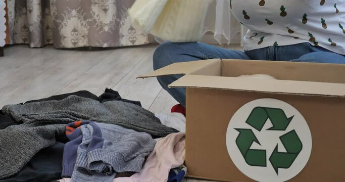 Sitting on the floor in the living room, a nice woman goes through old things. A woman puts old things in a box for donation, charity. Proper use of old clothes. Eco-friendly, recyclable.