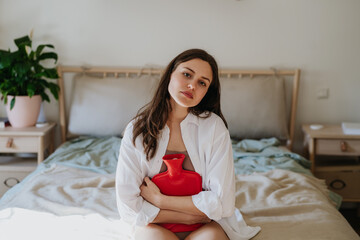 Woman at home suffering from menstrual pain, having cramps. Woman warming lower abdomen with a hot water bottle, endometriosis, and conditions causing pain in tummy.