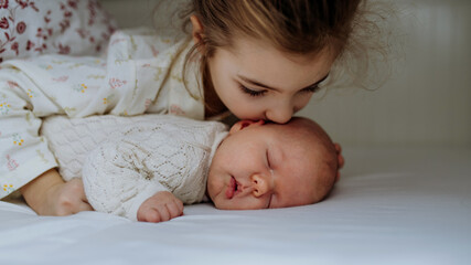 Portrait of big sister cuddling newborn, little baby. Girl kissing her sleeping new sibling in bed....