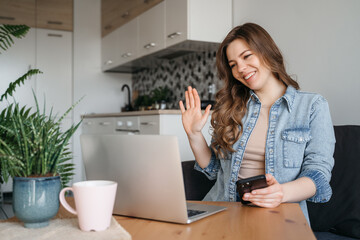 A woman sits at a table with laptop, cell phone, and houseplant, smiling