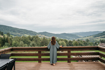 Young woman staying on terrace of wooden modern house with panoramic windows near pine forest. Concept of solitude and recreation on nature. Wellness and mindful resort. Beautiful place for vacation.