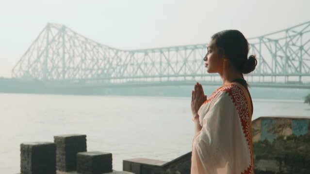 A young beautiful traditional Indian Asian woman or female Hindu devotee in an ethnic saree standing and making Namaste gesture on the riverbank praying or worshipping the Holy Ganges river