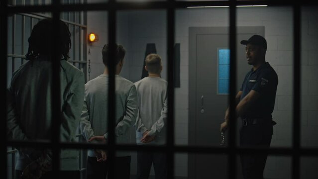 Group of prisoners stand near jail cell, warden talks with them. Multicultural teenagers serve imprisonment term in detention center. Young inmates in prison. Justice system. View through metal bars.