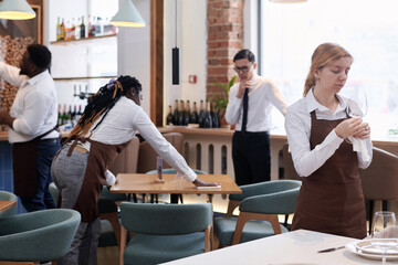 Medium long selective focus shot of ethnically diverse restaurant workers cleaning furniture and polishing glassware before opening
