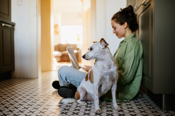 Young student e-learning on laptop at home, cute small dog wants to play. Businesswoman working...