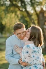 Fototapeta premium Beautiful woman kissing and hugging man stands on green grass at sunset. Happy couple in love embraces and kisses on field in sunset background. Female and male in a park on summer day. Photo closeup.