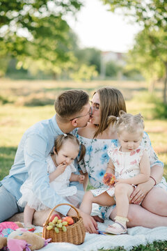 Portrait of mom, dad, two daughter. Young family eating food in park on vacation together. Concept of family holiday. Father, mother, baby girls sitting on green grass, picnic in nature. Couple kisses