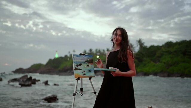 Artist paints seascape on canvas beside waves at sunset. Creative woman in black dress works on stormy beach art. Inspirational activity at ocean, dusk light. Hobby, leisure on coast. Slow motion.