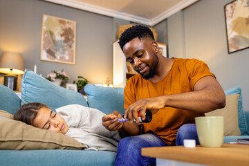 African American father takes care of his sick daughter at home and gives her syrup for temperature.