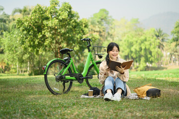 Peaceful young woman relaxing on green grass and reading book during summer day