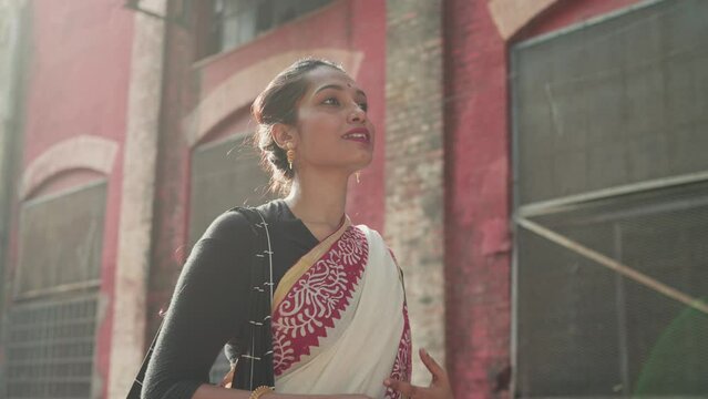 A young beautiful smiling Indian Asian hindu woman or female wearing traditional ethnic white saree is walking outdoors next to an old building and looking at a distance on sunny morning