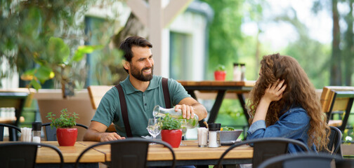 Young couple at date in restaurant, sitting on restaurant terrace. Boyfriend pouring water in...