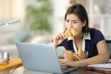 Stressed student eating a lot of bakery checking laptop