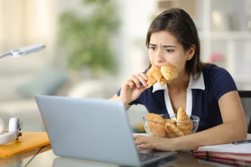 Fotobehang Stressed student eating a lot of bakery checking laptop © PheelingsMedia
