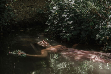 young woman floats gently in the waters of a river, wearing a dress with flowers and leaves. Serene...