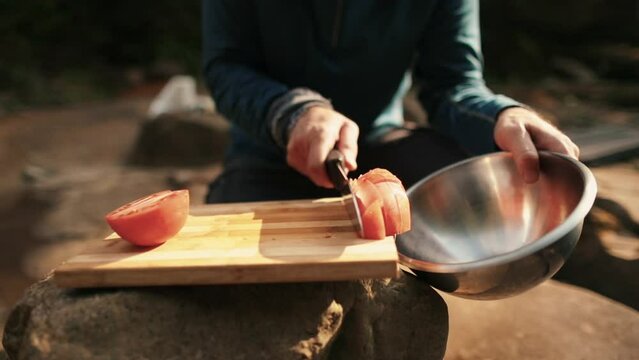 A man prepares tomatoes for consumption by dropping chopped pieces into a metal bowl outdoors on a hiking trip.