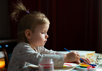 A young baby artist painting with watercolors. Adorable blonde girl of 2,5 years old learns to paint sitting at the table at home. The concept of early creative development of children.