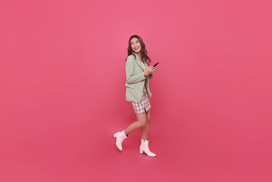 Young smiling happy asian woman 20s holding mobile cell phone and walking isolated on pink background. portrait People lifestyle concep