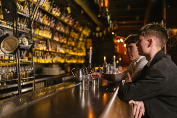 Guests drink beverages and talk sitting by candlelight at bar counter. Young man with dark haired friend meet in bar with subdued light - obrazy, fototapety, plakaty
