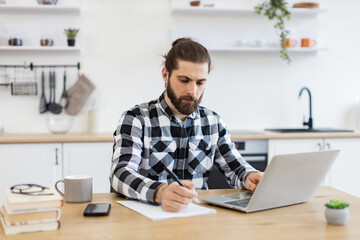 Handsome Caucasian man wearing checkered shirt writing on document while staying at home during midday. Efficient business owner improving contract while checking data on portable computer.
