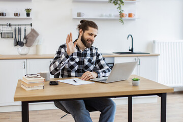 Cheerful bearded adult waving hello at webcam of portable computer while greeting colleague in home office. Proficient Caucasian consultant receiving online conference call using technologies.