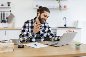 Plakaty  Proficient Caucasian consultant receiving online conference call using technologies. Cheerful bearded adult looking at webcam of portable computer while greeting colleague in home office.