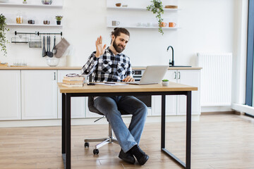 Cheerful bearded adult waving hello at webcam of portable computer while greeting colleague in home...