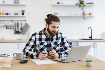 Young Caucasian man in wireless headset conducting online meeting with teammates via laptop in kitchen interior. Serious freelance worker showing effective communication ability during web conference.