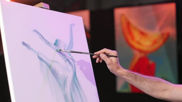 Close up view of inspired artist hand holding paintbrush and painting blue female ballet figure on canva indoors. Background picture in orange shades. Concept of art and creative.