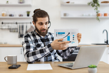 Attractive male holding paper with charts while speaking during online meeting via computer at home. Qualified data analyst demonstrating information about business growth in kitchen interior.
