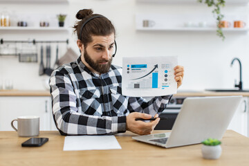 Qualified data analyst demonstrating information about business growth in kitchen interior. Attractive male holding paper with charts while speaking during online meeting via computer at home.