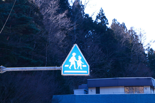 School roadside warning sign mounted on brown steel beam. Mountains, blue sky and bright sunlight in background. Symbol aluminum white, blue post about pedestrian crossing road sign traffic.