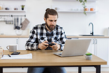 Portrait of young Caucasian man using wireless joystick and laptop sitting at wooden table at...