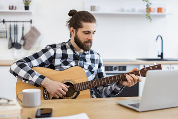 Cheerful Caucasian person holding string instrument while smiling at portable computer webcam in spacious apartment. Happy male teaching online guitar lesson using modern technologies.