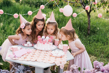 Summer outdoor kids birthday party. Group of happy Children celebrating birthday in park. Children blow candles on birthday cake. Kids party pink pastel decoration and food. Presents and sweets. - 761426813