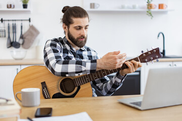 Happy male teaching online guitar lesson using modern technologies. Cheerful Caucasian person holding string instrument while smiling at portable computer webcam in spacious apartment. - obrazy, fototapety, plakaty