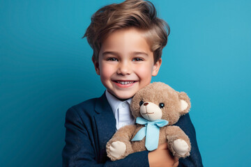 An adorable kid model in a suit, holding a teddy bear and smiling warmly against a captivating blue wall background.