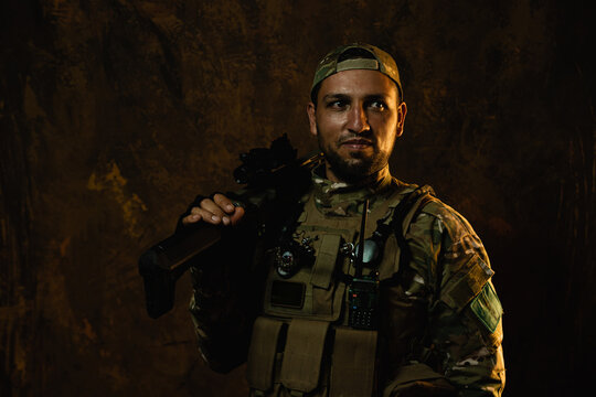 A Latin American soldier with a confident gaze stands with his rifle, outfitted in tactical combat gear