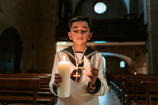 A solemn young boy, dressed in traditional First Communion garb, holds a lit candle and cross in a serene church setting